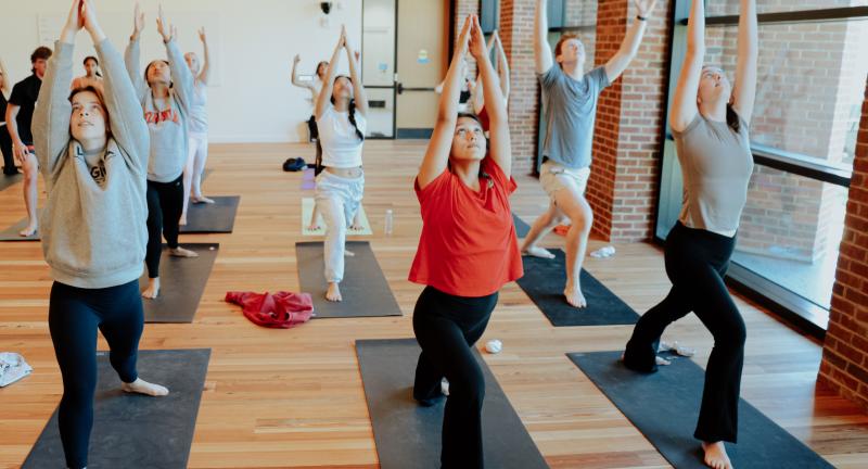 Students practice Ashtanga Yoga, reaching their arms up towards the ceiling in a room with wooden floors